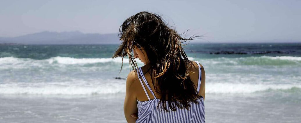 Eine junge Frau in weißem Kleid steht verträumt am Meer und blickt in die Ferne.