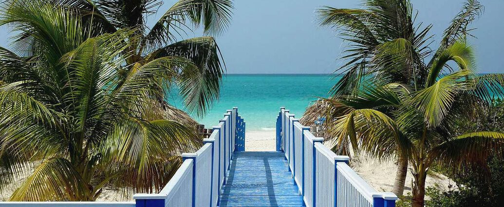 Ein kleiner weißer Steg führt direkt zu einem Traumstrand mit weißem Sand, Palmen und herrlich blauem Wasser.