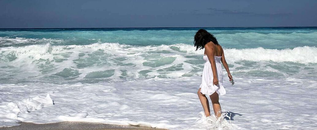 Eine junge Frau in einem weißen Kleid steht mit den Füßen im Meer am Strand.