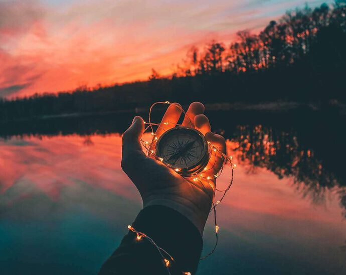 Eine Hand hält einen Kompass, die Umgebung dämmert in der Abendsonne.