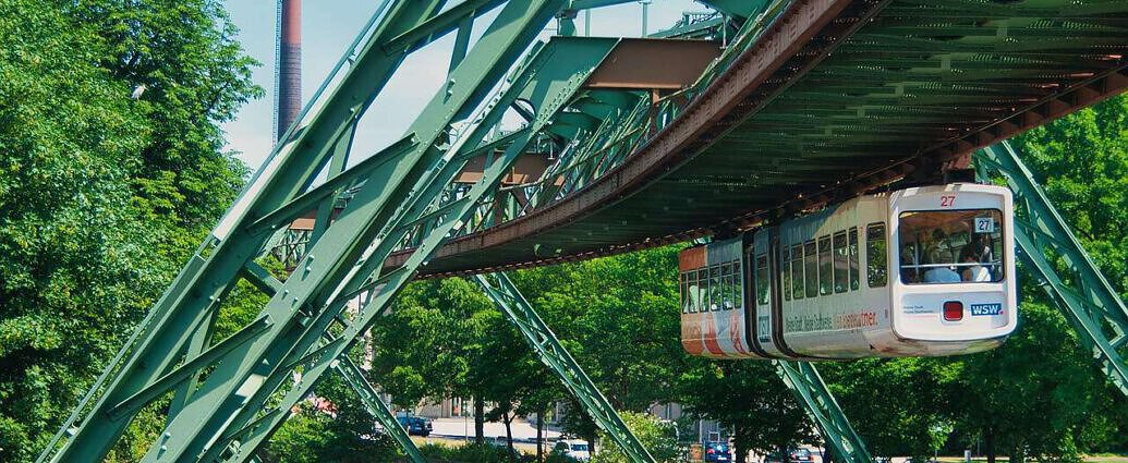 Ein Bild der Wuppertaler Schwebebahn, mit schöner Umgebung und Wupper, unter der ein Fluss zu sehen ist.