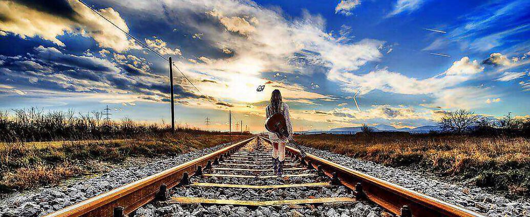 Eine junge Frau, die eine Gitarre auf dem Rücken trägt, wandert auf einer Eisenbahnstrecke entlang.