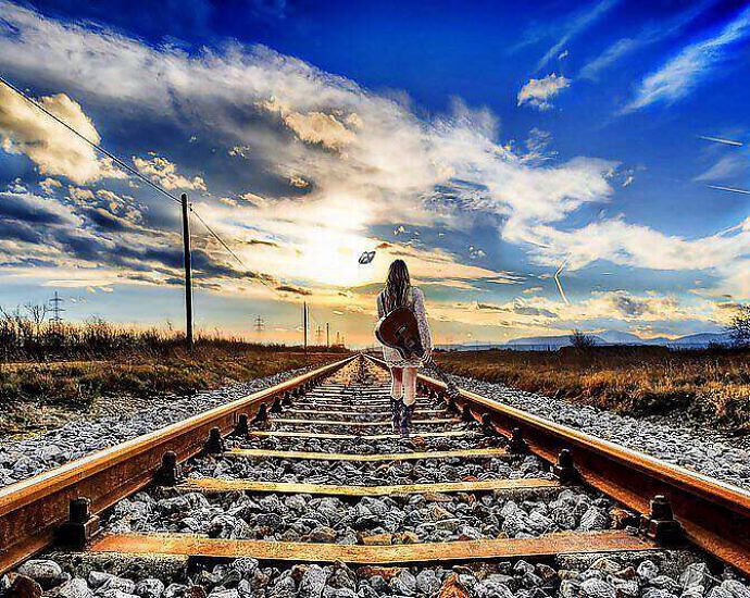 Eine junge Frau, die eine Gitarre auf dem Rücken trägt, wandert auf einer Eisenbahnstrecke entlang.