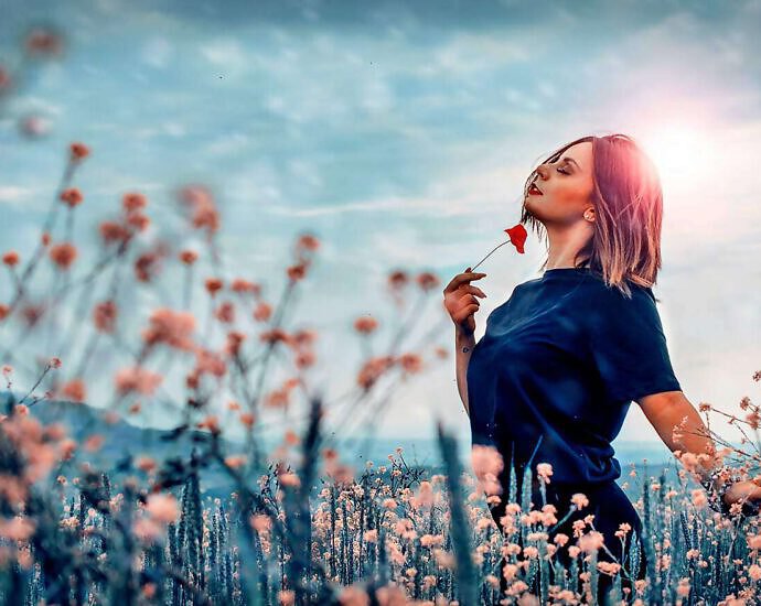 Eine junge Frau mit langen braunen Haaren steht auf einer Blumenwiese und blickt verträumt in den Himmel.