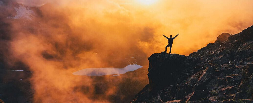 Ein Mann streckt seine Arme auf einem hohen Berg aus. Im Hintergrund geht gerade die Sonne auf.
