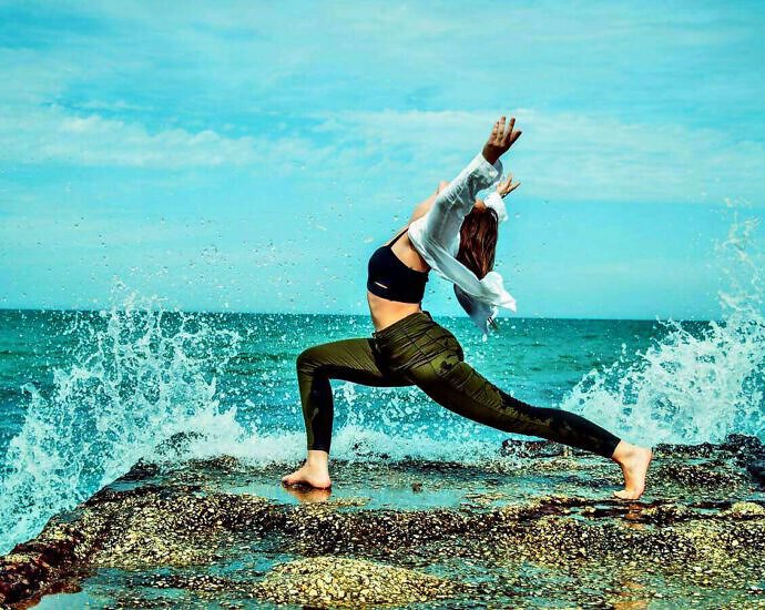 Eine junge Frau in grünen Jeans verrenkt ihren Körper in einer Pose der Freude direkt am Meer.