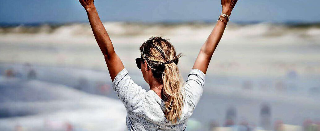 Eine junge Frau hebt die Arme, sie steht am Meer.