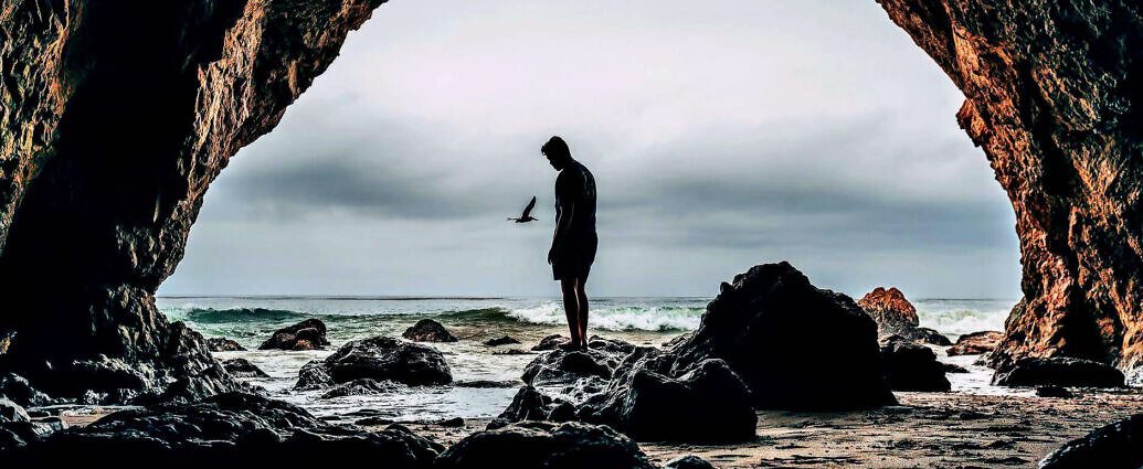 Ein Mann steht verträumt am Meer, vor ihm ein Felsbogen.