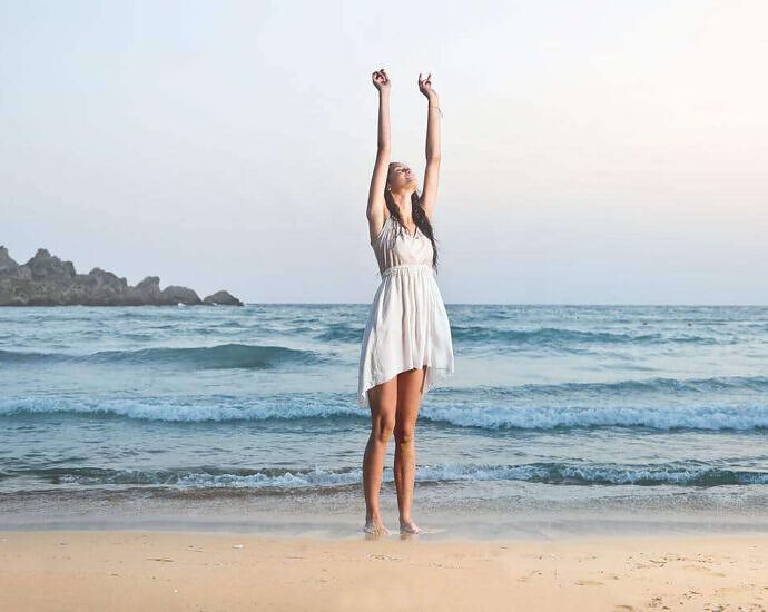 Eine junge Frau steht mit den Füßen im Wasser am Strand, trägt ein weißes Kleid und hebt vor Freude die Arme hoch.