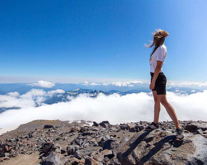 Eine junge Frau steht an einem Berghang und blickt auf die Berge vor ihr.