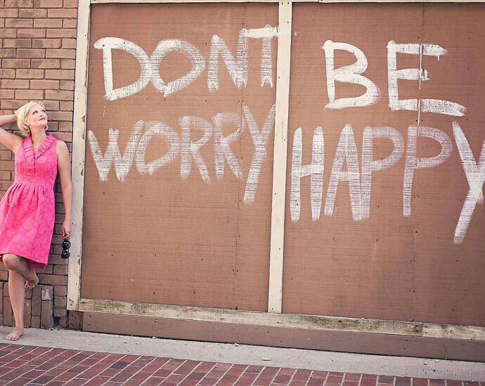Eine junge blonde Frau in einem rosa Kleid steht an einer Wand mit der Aufschrift Dont Worry Be Happy.