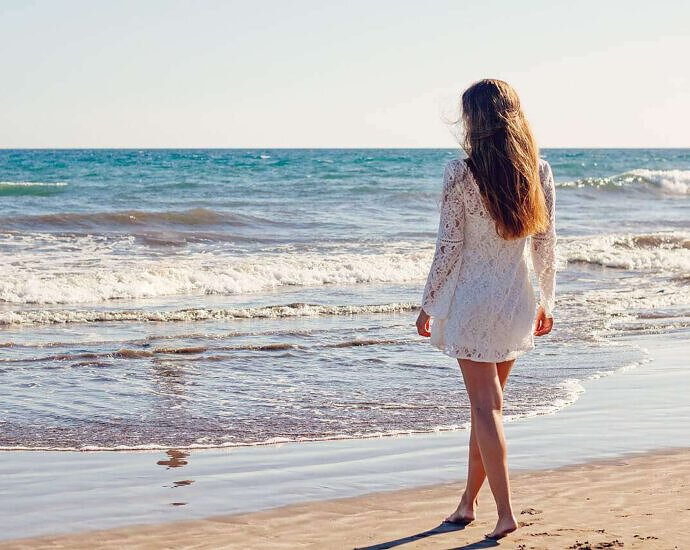 Am Strand steht eine Frau im weißen Strandkleid und schaut auf das Meer.