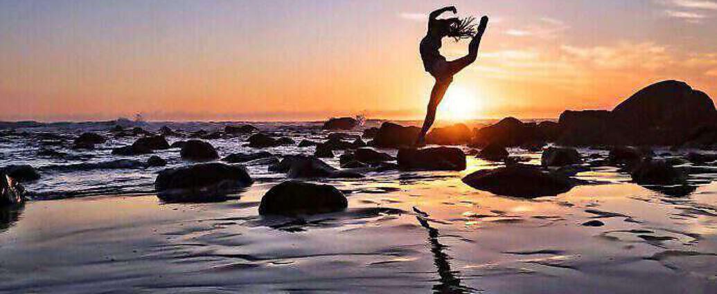 Eine junge Frau springt vor Freude im Sonnenuntergang am Strand in die Luft.