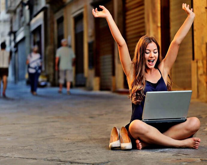 Eine junge Frau sitzt auf dem Boden. Auf ihrem Schoß liegt ein Laptop. Vor Freude reißt sie die Arme hoch.