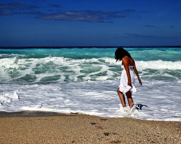 Eine junge Frau im weißen Kleid spielt mit den Füßen im Meer am Strand.