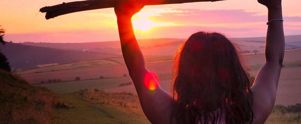 Eine junge Frau hält bei Sonnenuntergang einen Stock mit beiden Armen in die Luft.