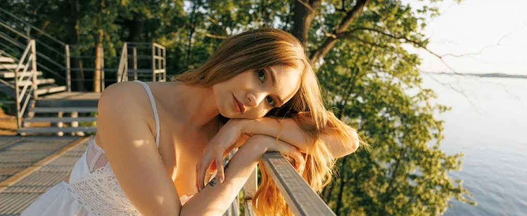 Eine Frau im weißen Kleid lehnt an einem Zaun, neben ihr ein See.