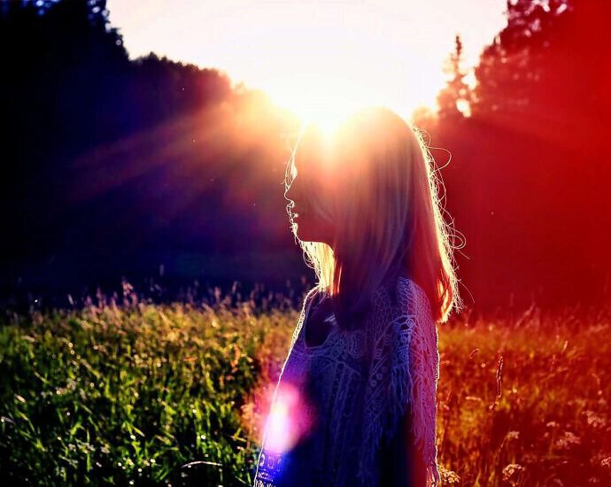 Zu sehen ist die Silhouette einer jungen Frau, die entspannt am Rande eines Waldes in der Abendsonne steht.