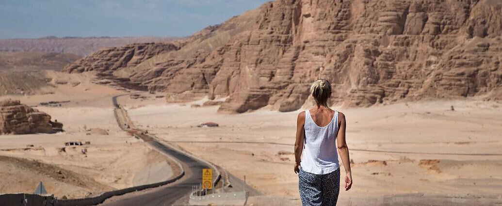 Ein Bild von einer Frau, die auf einem Highway steht und geradeaus schaut. Vor ihr sind Berge zu sehen.