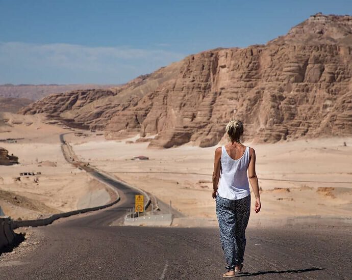 Ein Bild von einer Frau, die auf einem Highway steht und geradeaus schaut. Vor ihr sind Berge zu sehen.