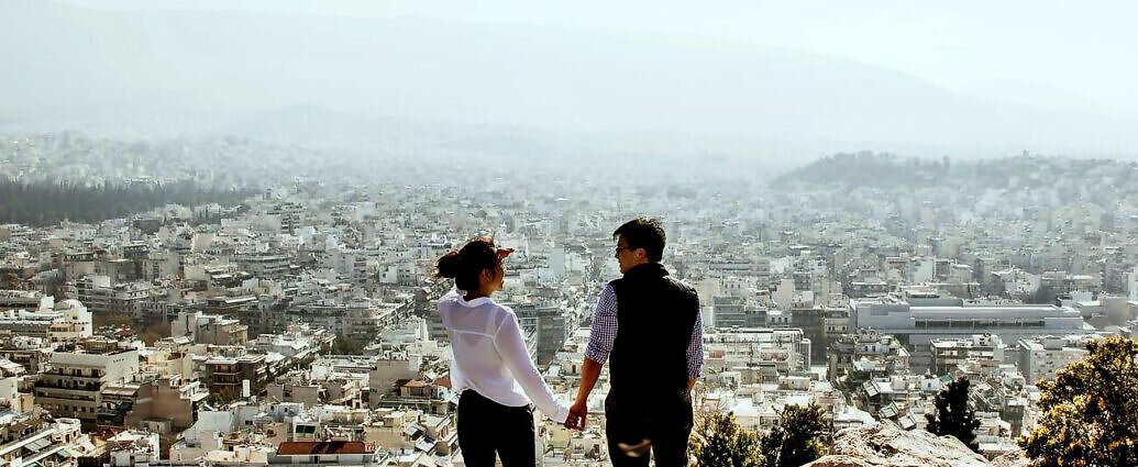 Ein junges Paar schaut Hand in Hand von einer Anhöhe auf die Stadt unter ihnen.