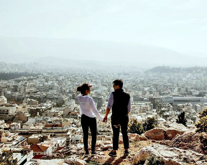 Ein junges Paar schaut Hand in Hand von einer Anhöhe auf die Stadt unter ihnen.