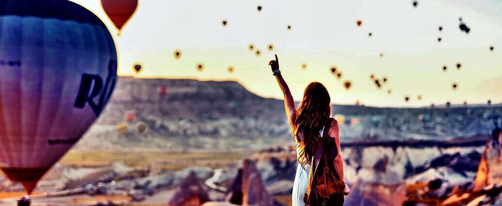 Eine junge Frau in einem weißen Kleid steht am Berghang und winkt den Ballonfahrern zu.