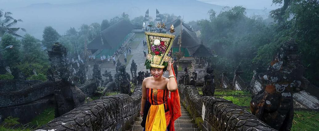 Das Bild zeigt eine junge Frau in traditioneller Kleidung auf Bali, Indonesien. Im Hintergrund ist der Dschungel zu sehen.