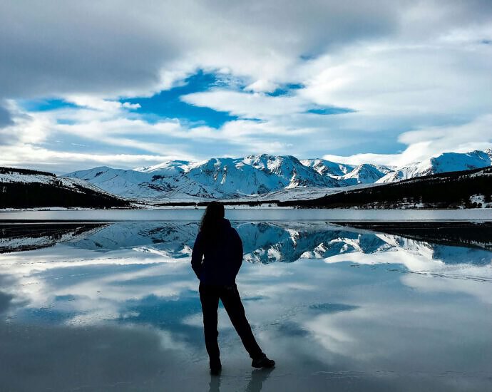 Eine junge Frau steht auf einem See in Patagonien und blickt auf die schneebedeckten Berge.