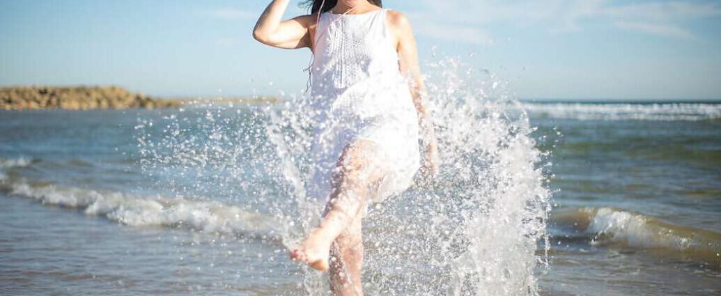 Eine Frau, die lachend am Ufer des Meeres steht und mit den Füßen im Wasser planscht.