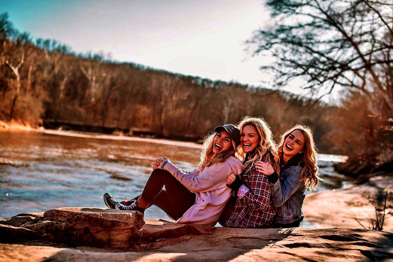 Drei junge Frauen sitzen hintereinander und lachen, vor ihnen ein Fluss.