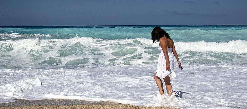 Eine Frau in weißem Kleid steht am Strand, die Füße im Wasser der Wellen.