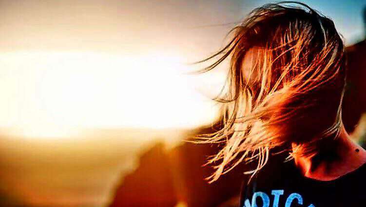 Eine junge Frau schaut verträumt in den Sonnenaufgang, ihr Haar weht leicht im Wind.