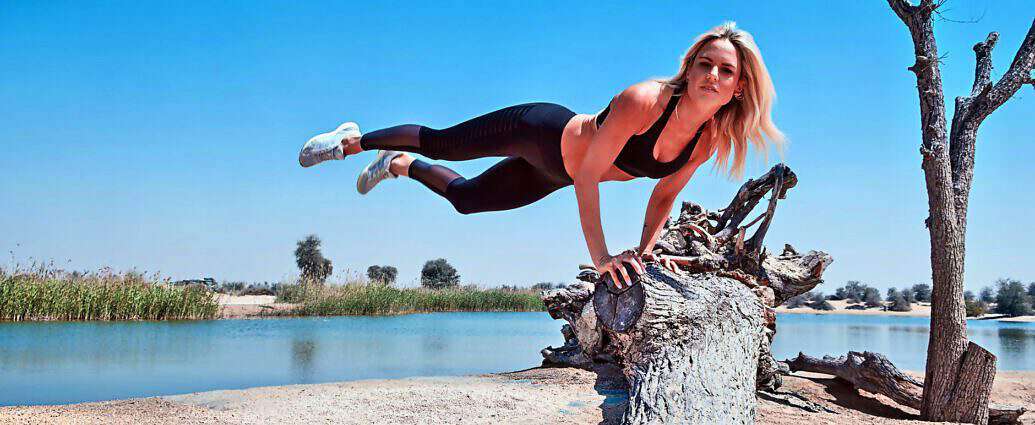 Eine junge Frau in enger Trainingskleidung springt seitlich an einem Baumstamm hoch.