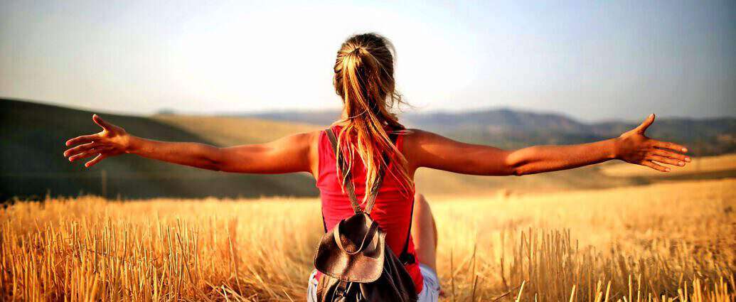 Eine junge Frau sitzt in einem Strohfeld, blickt geradeaus und streckt die Arme der Sonne entgegen.