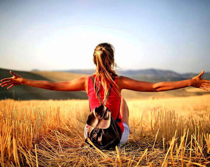 Eine junge Frau sitzt in einem Strohfeld, blickt geradeaus und streckt die Arme der Sonne entgegen.