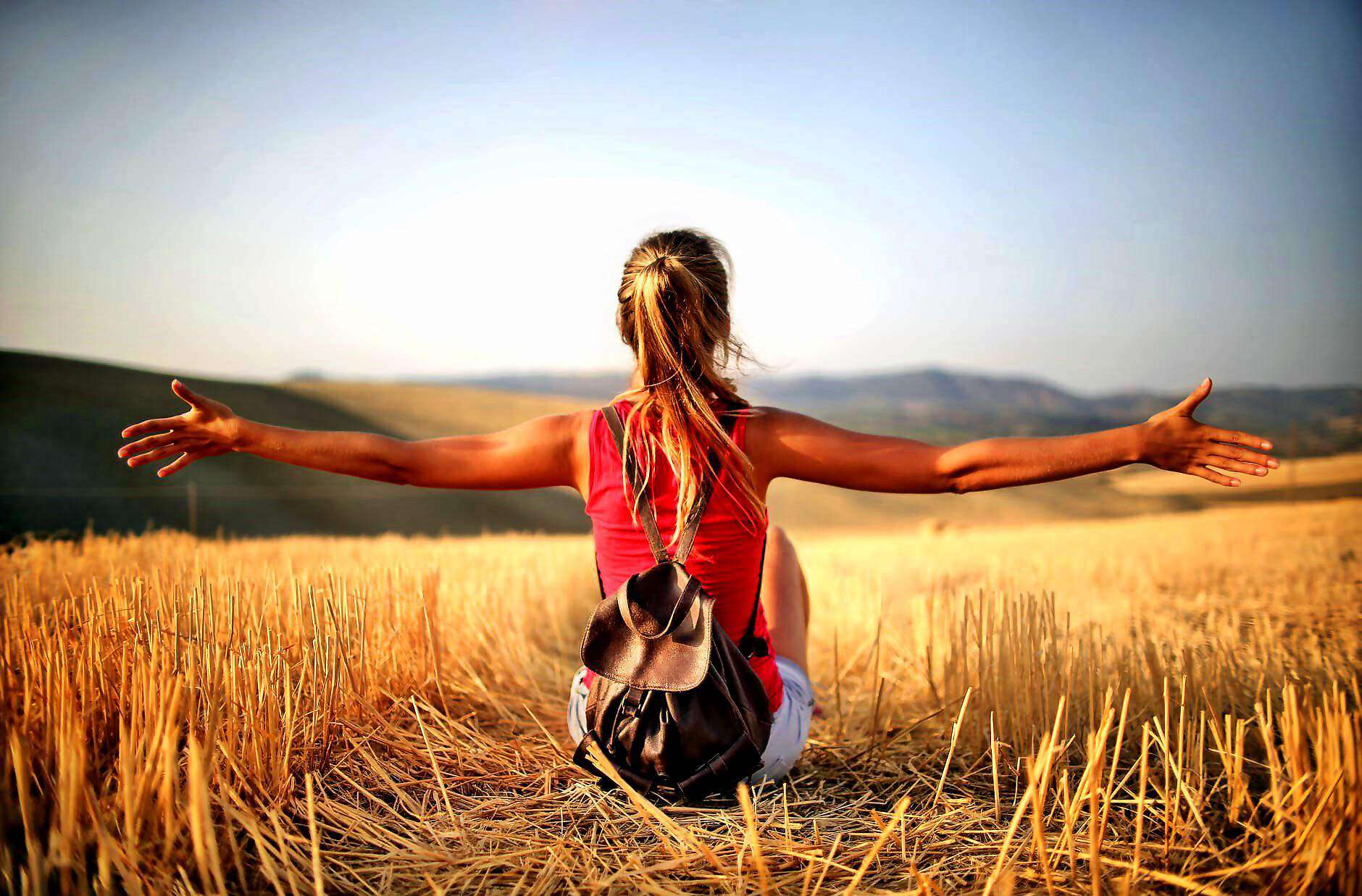 Eine junge Frau sitzt in einem Strohfeld, blickt geradeaus und streckt die Arme der Sonne entgegen.