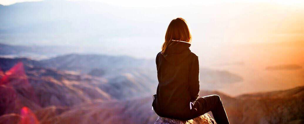 Eine junge Frau sitzt am Rande eines Berghanges und blickt in die Ferne.