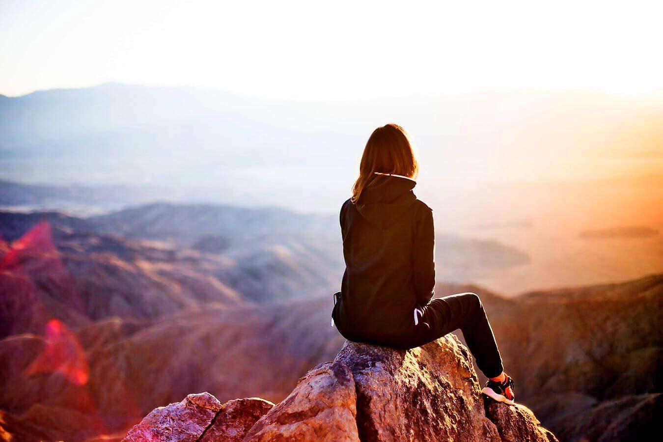 Eine junge Frau sitzt am Rande eines Berghanges und blickt in die Ferne.