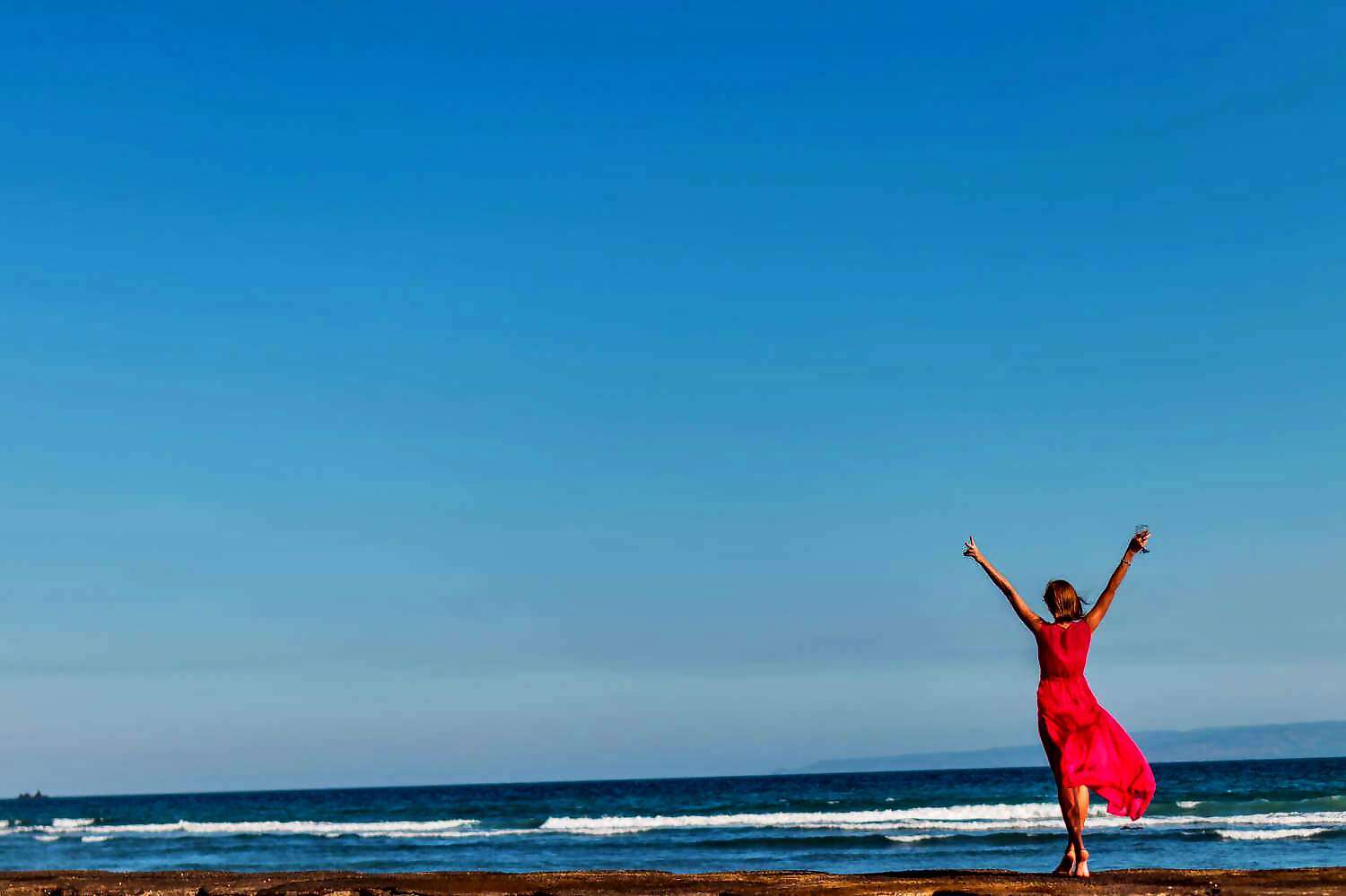 Eine Frau in rotem Kleid hebt freudig die Arme und steht am Meer.