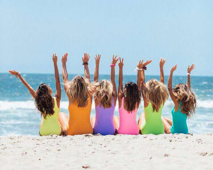 Junge Frauen sitzen am Strand und reißen vor Freude die Arme in die Höhe.