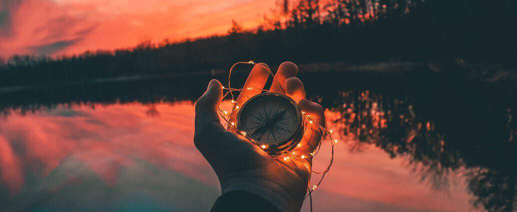 Eine Hand hält einen Kompass, die Umgebung dämmert in der Abendsonne.