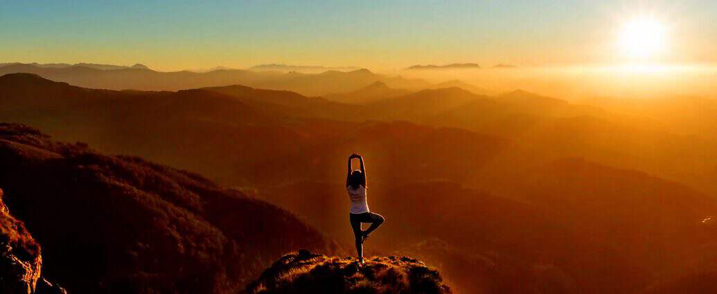 Eine junge Frau steht auf einer Bergspitze in der Abendsonne und streckt sich vor Freude.