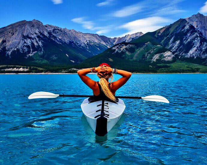 Eine junge Frau steht mit ihrem Kanu auf einem See und beobachtet die Berge vor sich.