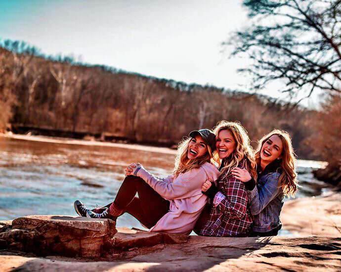 Drei junge Frauen, die nacheinander am Fluss sitzen und lachen, strahlen Selbstvertrauen aus.