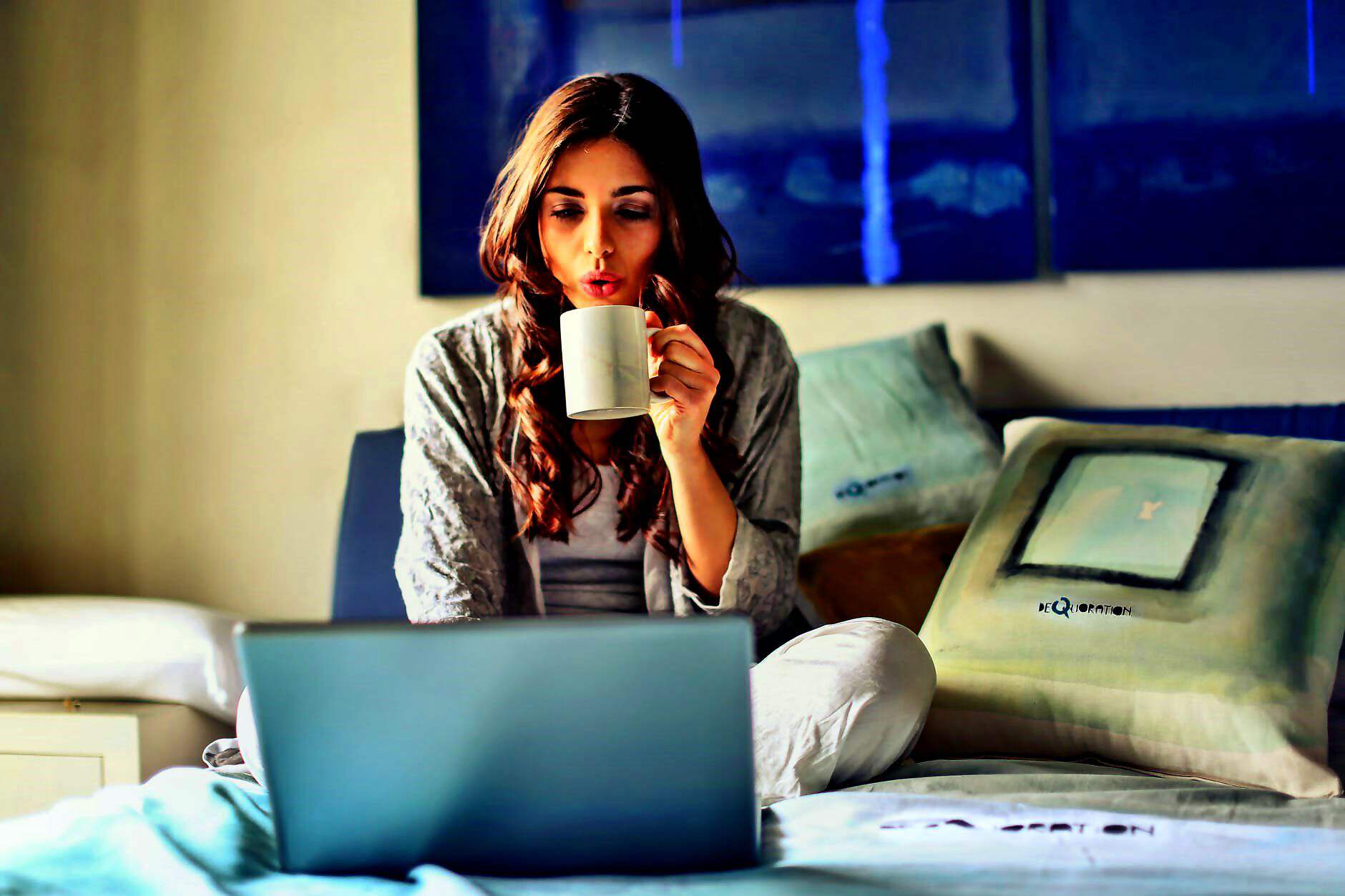 Eine junge Frau sitzt im Schneidersitz auf dem Bett und schaut gespannt auf den Laptop. In der Hand hält sie einen Becher Kaffee.