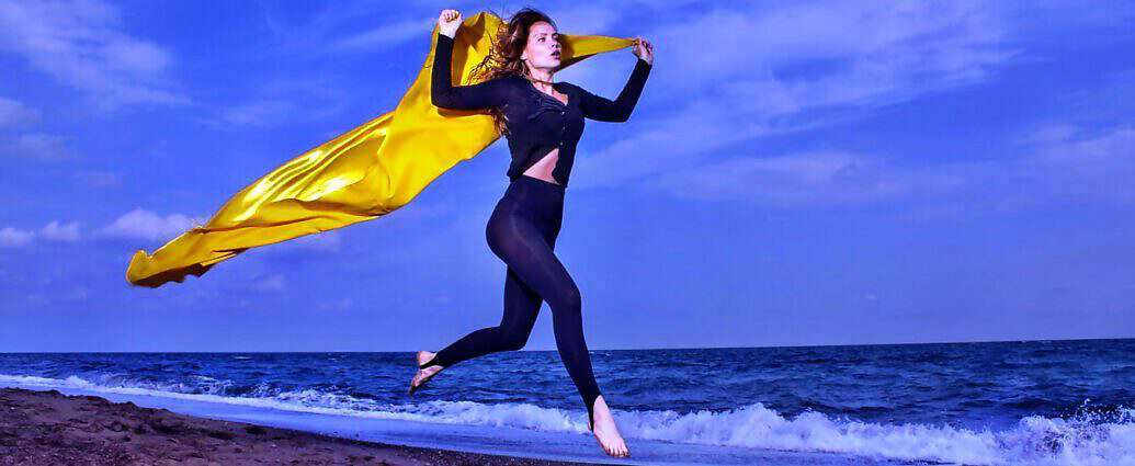 Eine junge Frau in schwarzer Leggings und mit einem gelben Tuch, das in der Luft wirbelt, springt vor Freude am Strand in die Höhe.