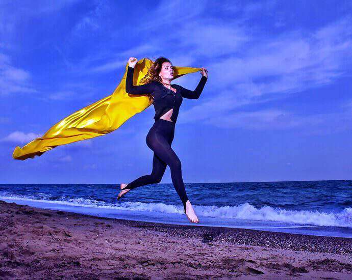 Eine junge Frau in schwarzer Leggings und mit einem gelben Tuch, das in der Luft wirbelt, springt vor Freude am Strand in die Höhe.