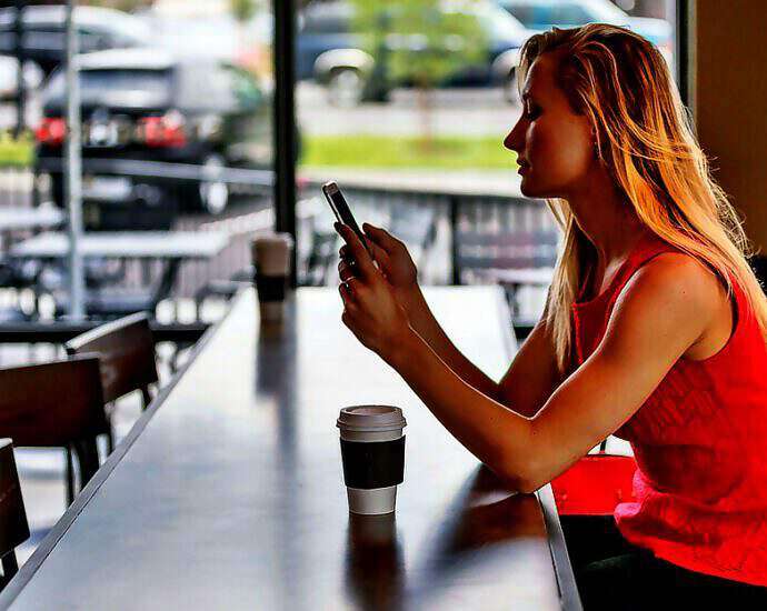 Eine junge blonde Frau sitzt in einem Café. Vor ihr steht ein Pappbecher mit Kaffee. Sie schaut auf ihr Smartphone.