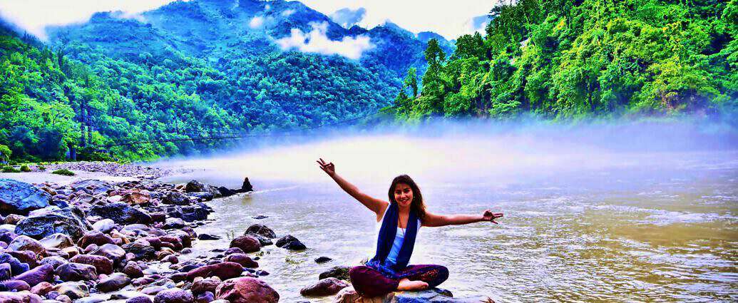 In einer wunderschönen Landschaft sitzt eine junge Frau im Schneidersitz am Ufer eines Flusses.